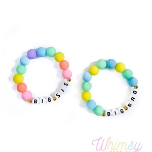 May include: Two colorful beaded bracelets. One bracelet spells "BIG SIS" and the other spells "BIG BRO" with white letter beads. The bracelets feature pastel-colored beads in a rainbow pattern, with gold accent beads.