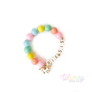 May include: A colorful beaded bracelet with pastel-colored beads in shades of pink, blue, yellow, and green. The bracelet features white letter beads spelling "BIG SIS" and a gold unicorn charm. The bracelet is designed for children.
