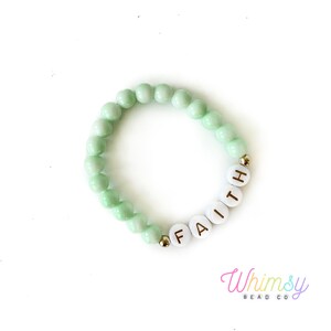 May include: A light green beaded bracelet with the word "FAITH" spelled out in white letter beads. The bracelet has gold-colored accent beads and is on a white background. The Whimsy Bead Co. logo is in the bottom right corner.