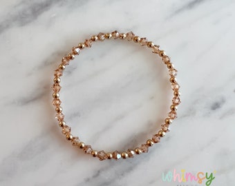 May include: A delicate bracelet featuring small, faceted, light brown beads interspersed with gold-colored metallic beads. The bracelet is circular and sits on a white and gray marble surface. The Whimsy Bead Co. logo is visible in the bottom right corner.