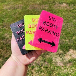 May include: Three rectangular signs with the text "BIG BOOTY PARKING" in black. The signs are black, neon yellow, and neon pink. The pink sign has a black arrow pointing left and right.