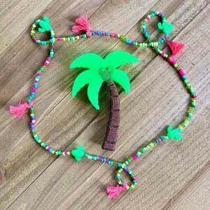 Può includere: Una collana di perline a tema tropicale. Presenta un ciondolo a forma di palma verde brillante e piccoli ciondoli a forma di cactus verde. Le perline sono rosa, blu e verdi, con nappe rosa.
