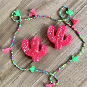 Puede incluir: Un vibrante conjunto de collar y pendientes con temática de cactus. Los pendientes son de color rosa brillante con purpurina, y el collar tiene cuentas coloridas, abalorios de cactus verdes y borlas rosas. El conjunto se exhibe sobre una superficie de madera.