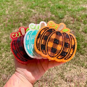 Op de afbeelding: Een verzameling pompoenvormige decoraties in rood, wit, teal en oranje. De pompoenen hebben glitteraccenten en ruitpatronen. Elke pompoen heeft een krullende stengel en het woord "cel" erop geschreven. Perfect voor de herfst.