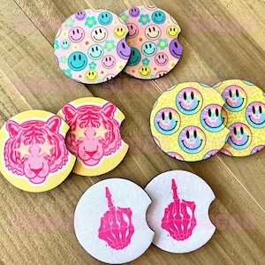 May include: A collection of car coasters with various designs. The coasters feature smiley faces, tiger faces, and a hand giving the middle finger. Colors include pink, yellow, blue, and purple. The coasters are round with a small notch.