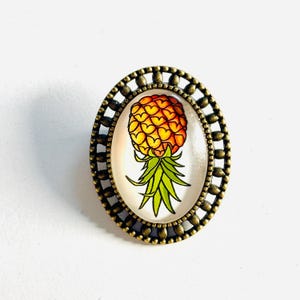 May include: An oval brooch featuring a central illustration of a pineapple with orange and yellow heart-shaped segments and green leaves. The artwork is encased in a decorative, antique bronze-toned metal frame with a scalloped, beaded edge.