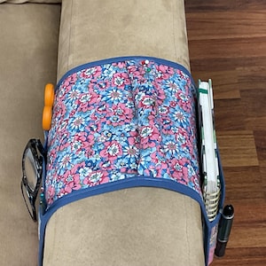 May include: A floral print couch arm organizer with blue trim, holding scissors, glasses, a notebook, and a pen. The organizer is wrapped around the arm of a beige couch. The floral pattern includes pink and blue flowers.