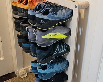 May include: A wooden shoe rack with nine pairs of trainers. The shoes are a mix of black, blue, and grey colours. The rack is mounted on a white wall.
