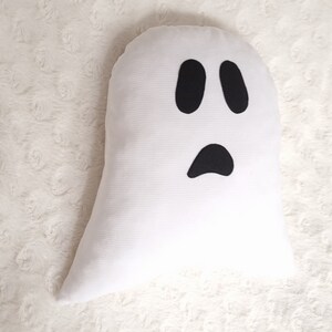 May include: White ghost pillow with black felt eyes and mouth. The pillow is shaped like a ghost and has a simple, friendly expression.