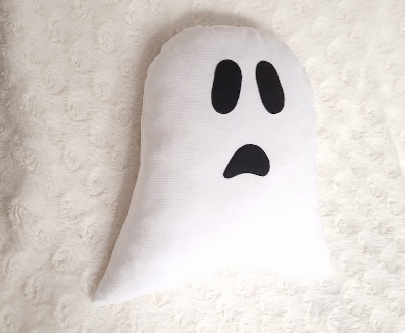 May include: White ghost pillow with black felt eyes and mouth. The pillow is shaped like a ghost and has a simple, friendly expression.
