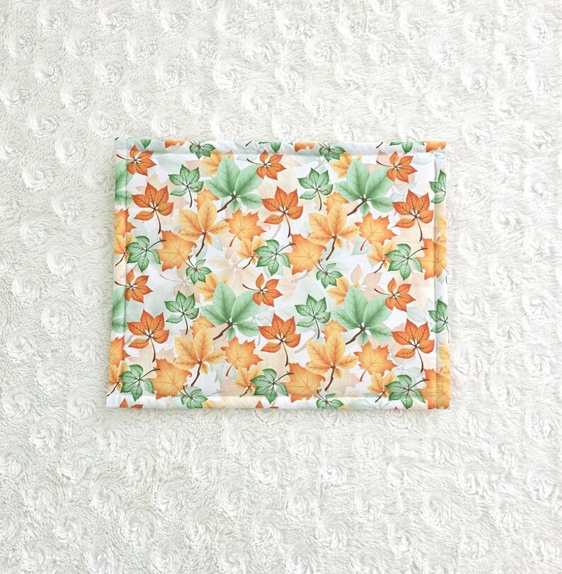 Op de afbeelding: Rechthoekig stoffen item met een herhaald patroon van oranje, groene en gele herfstbladeren op een witte achtergrond. De randen zijn afgezet met een lichtgroene stof.