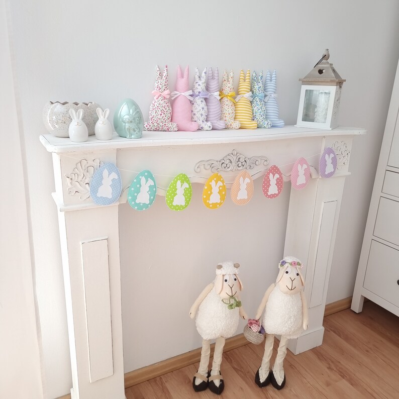 May include: A white mantelpiece decorated with a pastel Easter bunny garland and a string of pastel egg-shaped ornaments with white bunny silhouettes. Two white fluffy sheep figurines stand in front of the mantelpiece.