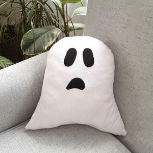May include: White ghost-shaped pillow with black felt eyes and a mouth. The pillow is sitting on a gray couch.