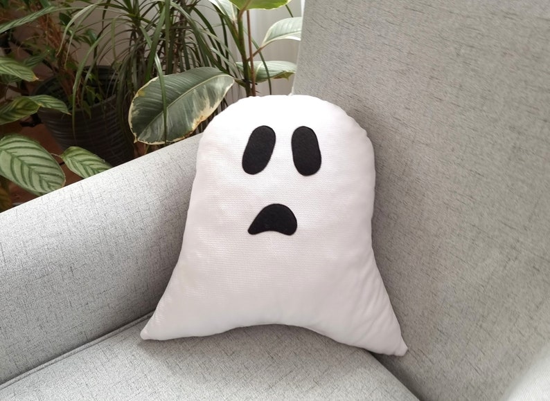 May include: White ghost-shaped pillow with black felt eyes and a mouth. The pillow is sitting on a gray couch.