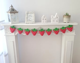 May include: A white mantelpiece with a garland of red and white felt strawberries with green leaves. The garland is hung above a white sign that says "You are our" and a white ceramic house.