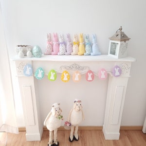 May include: A white mantelpiece with a pastel bunny garland and two white ceramic lambs. The garland is made of pastel colored eggs with white bunny silhouettes. The garland is hung above the mantelpiece.