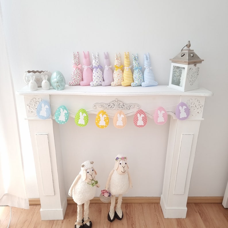 May include: A white mantelpiece with a pastel bunny garland and two white ceramic lambs. The garland is made of pastel colored eggs with white bunny silhouettes. The garland is hung above the mantelpiece.