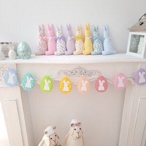 May include: A pastel colored Easter bunny garland with white bunny silhouettes on each egg. The garland is hanging on a white mantelpiece. There are also eight pastel colored fabric bunnies on the mantelpiece.