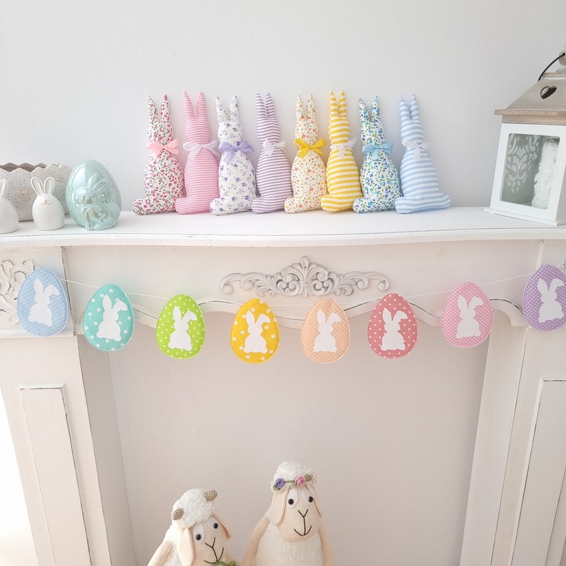 May include: A pastel colored Easter bunny garland with white bunny silhouettes on each egg. The garland is hanging on a white mantelpiece. There are also eight pastel colored fabric bunnies on the mantelpiece.
