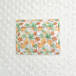 Op de afbeelding: Een vierkant etui met ritssluiting met een herhalend patroon van herfstbladeren in groen, oranje en geel op een witte achtergrond. De etui heeft een witte rits en een dunne groene rand.
