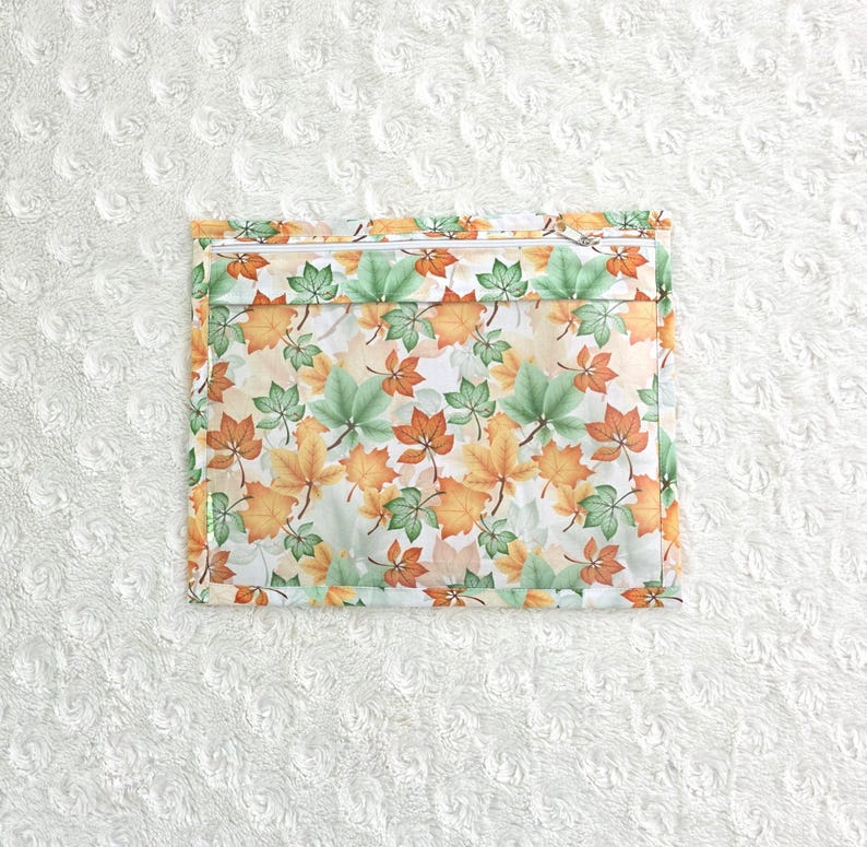 Op de afbeelding: Een vierkant etui met ritssluiting met een herhalend patroon van herfstbladeren in groen, oranje en geel op een witte achtergrond. De etui heeft een witte rits en een dunne groene rand.