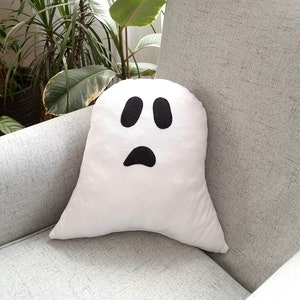May include: A white ghost-shaped pillow with black eyes and a frowning mouth. The pillow is sitting on a gray couch.