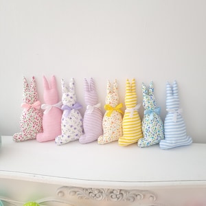 May include: Eight handmade fabric Easter bunny plush toys in a row. The bunnies are in various colors and patterns, including pink, purple, yellow, and blue. They are all made with a striped fabric and have a floral pattern.