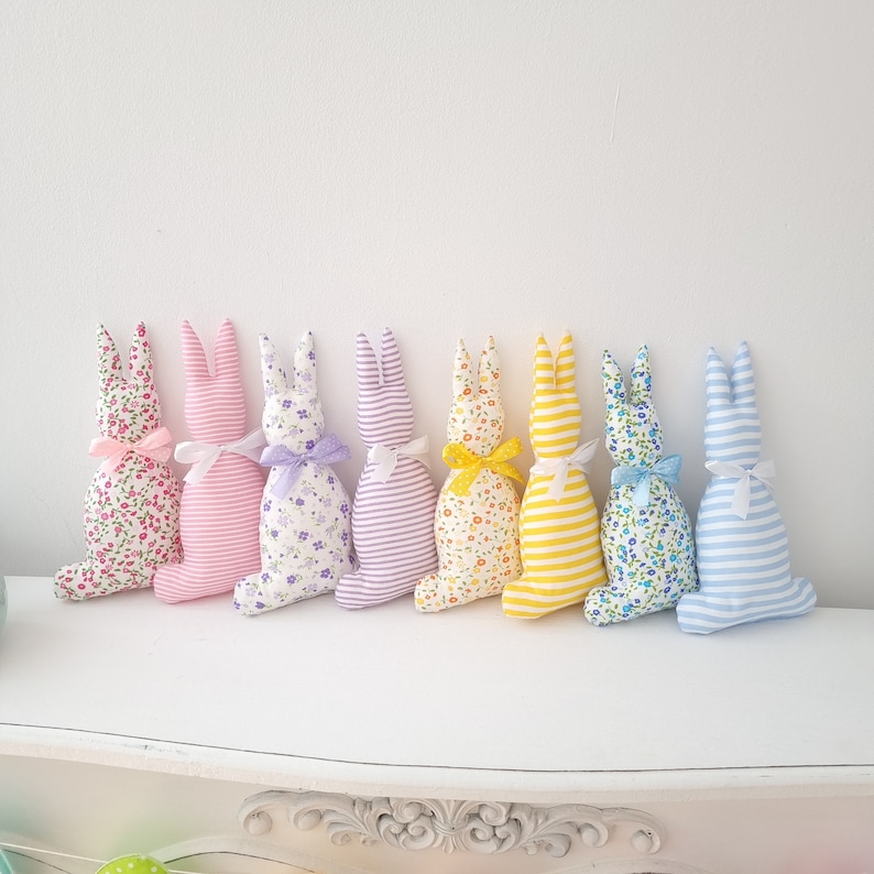 May include: Eight handmade fabric Easter bunny plush toys in a row. The bunnies are in various colors and patterns, including pink, purple, yellow, and blue. They are all made with a striped fabric and have a floral pattern.