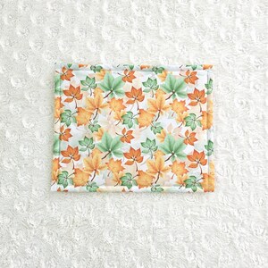 Op de afbeelding: Rechthoekig stoffen item met een herhaald patroon van oranje, groene en gele herfstbladeren op een witte achtergrond. De randen zijn afgezet met een lichtgroene stof.