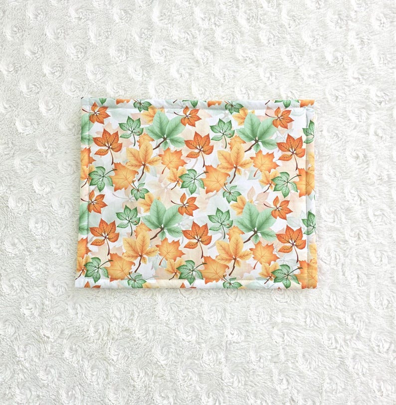Op de afbeelding: Rechthoekig stoffen item met een herhaald patroon van oranje, groene en gele herfstbladeren op een witte achtergrond. De randen zijn afgezet met een lichtgroene stof.