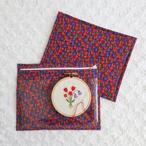 Op de afbeelding: Een rechthoekige projecttas met een doorzichtige vinyl voorkant en een achterkant van bloemenstof met rode en paarse tulpen op een donkere ondergrond. Binnenin zit een ronde houten borduurring met een geborduurd tulpenontwerp op stof.
