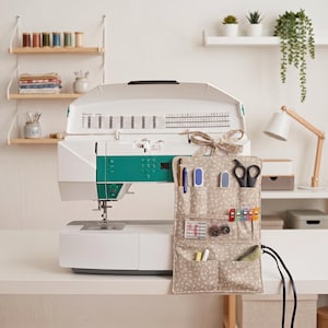 Può includere: Una macchina da cucire bianca e verde acqua con un organizer in tessuto floreale beige attaccato al lato. L'organizer presenta diverse tasche contenenti strumenti per cucire tra cui forbici, aghi, clip, un metro a nastro e penne.