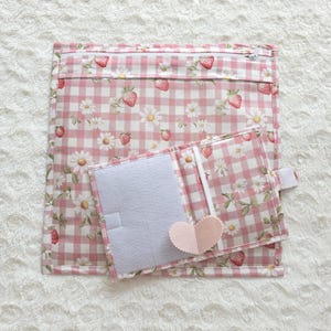 Könnte beinhalten: Eine quadratische Stofftasche mit Reißverschluss und ein kleineres passendes Stoffetui mit rosa-weißem Vichy-Muster, bedruckt mit Erdbeeren und weißen Gänseblümchen. Das Etui hat ein hellgraues Innenfutter und eine herzförmige Lasche.