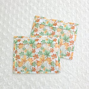 Op de afbeelding: Twee rechthoekige zakjes met ritssluiting met een herfstbladerenpatroon in groen, oranje en geel. De zakjes liggen op een wit, getextureerd oppervlak. De ritsen zijn wit.