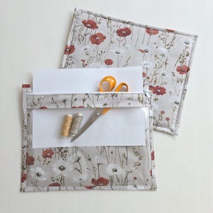 May include: Two rectangular fabric pouches featuring a beige background with a pattern of red and white poppies and thin green stems. The front pouch has a clear vinyl window, a silver zipper, and contains a white sheet of paper, orange scissors, and two small spools of thread.
