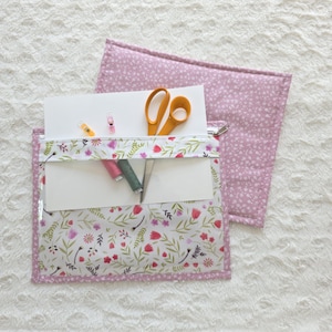Op de afbeelding: Een rechthoekig stoffen etui met een doorzichtig voorpaneel met een bloemenprint van rode, roze en groene botanische elementen. Het etui heeft een roze bloemige achterkant, een ritssluiting en bevat een gele schaar, twee garenklosjes en clips.