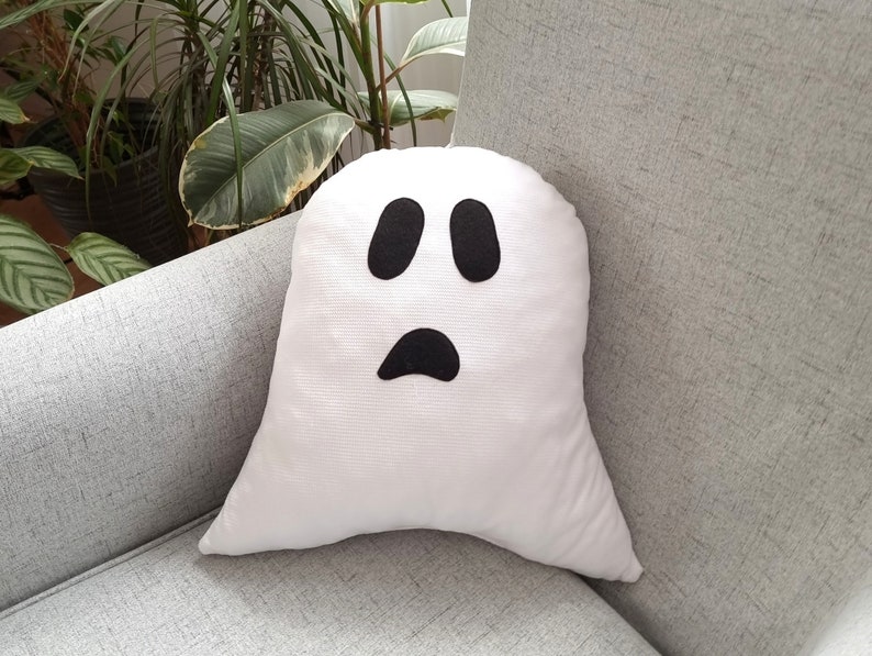 May include: A white ghost-shaped pillow with a black face and two eyes. The pillow is sitting on a gray couch.
