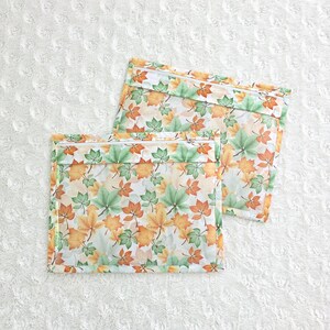 Op de afbeelding: Twee rechthoekige zakjes met ritssluiting met een herfstbladerenpatroon in groen, oranje en geel. De zakjes liggen op een wit, getextureerd oppervlak. De ritsen zijn wit.
