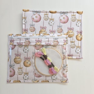 Può includere: Una borsa per progetti rettangolare in tessuto con un pannello frontale in vinile trasparente caratterizzato da un motivo ripetuto di palline di Natale rosa, oro e bianche con fiocchi. All'interno c'è un telaio da ricamo rotondo con tre matassine.