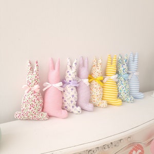 May include: Seven plush bunny rabbits in various colors and patterns. The bunnies are all facing the same direction and are arranged in a row. The bunnies are made of fabric and have long ears.