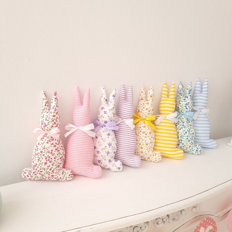 May include: Seven plush bunny rabbits in various colors and patterns. The bunnies are all facing the same direction and are arranged in a row. The bunnies are made of fabric and have long ears.