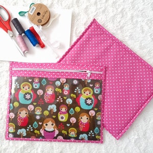 Può includere: Un cuscino rosa a pois e una pochette con cerniera abbinata con una finestra trasparente. La pochette presenta un motivo colorato di bambole russe, alberi e fiori. Sono visibili anche materiali da cucito, tra cui forbici e filo.