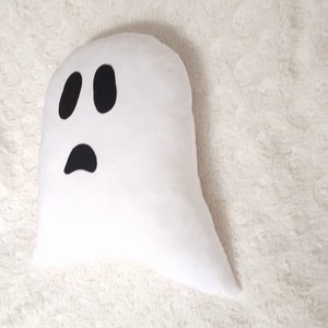 May include: White ghost-shaped pillow with a black felt face. The ghost has two large eyes and a small mouth.