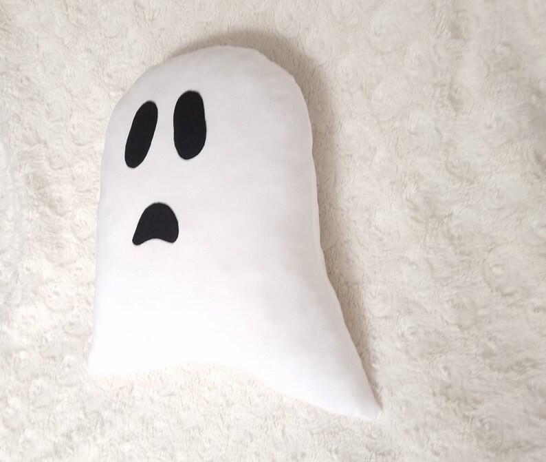 May include: White ghost-shaped pillow with a black felt face. The ghost has two large eyes and a small mouth.