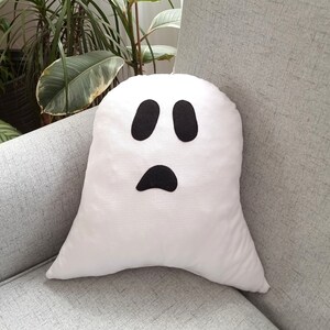 May include: A white ghost-shaped pillow with a black face and two eyes. The pillow is sitting on a gray couch.