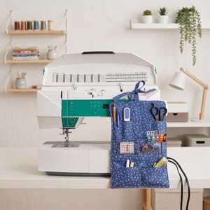 May include: A blue fabric sewing organizer with a small white floral pattern, featuring multiple pockets containing sewing tools like scissors, needles, clips, and a measuring tape, positioned in front of a white and teal sewing machine.