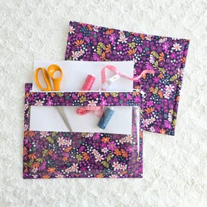 Puede incluir: Un organizador de costura con estampado floral con un bolsillo frontal transparente, que contiene tijeras, carretes de hilo y una cinta métrica. La tela presenta un fondo morado oscuro con flores coloridas. Una pieza de tela a juego está al lado.