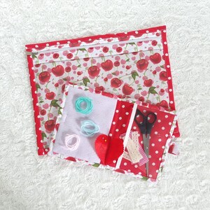 May include: A red and white fabric zippered pouch with a cherry print. The pouch has a smaller, clear pouch attached to it. The smaller pouch has a red heart, a small piece of lace, and a pair of scissors inside.
