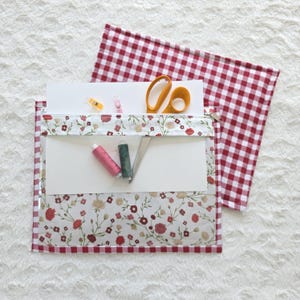 Puede incluir: Un estuche rectangular para proyectos con panel frontal transparente de tela blanca con estampado floral rojo y rosa. El estuche tiene un borde de cuadros vichy rojos y blancos, cierre de cremallera, tijeras amarillas, dos carretes y clips.