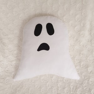 May include: White ghost-shaped pillow with a black face and two eyes. The pillow is on a white fuzzy surface.